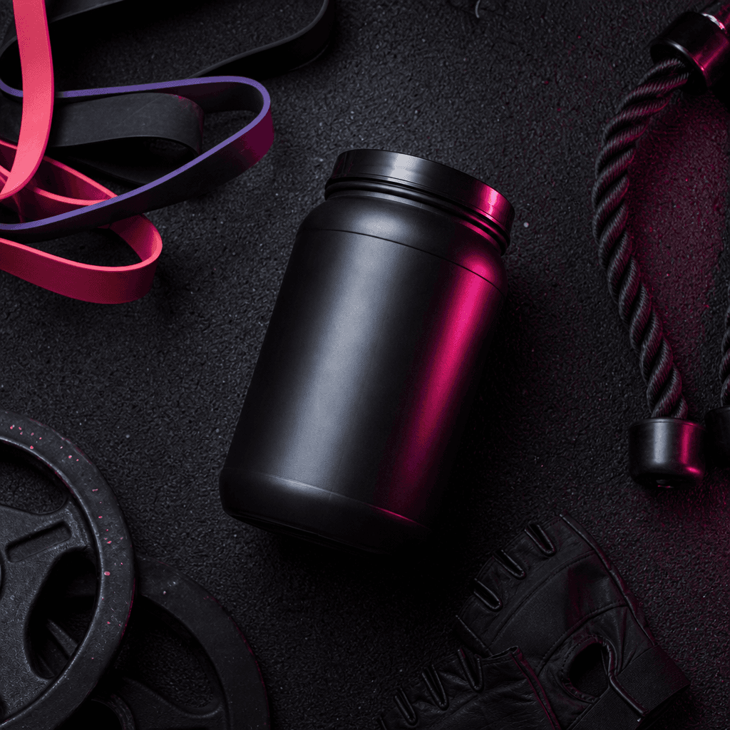 Overhead flat-lay of a premium sports supplement canister, resistance bands, and gym gloves arranged on black rubber flo