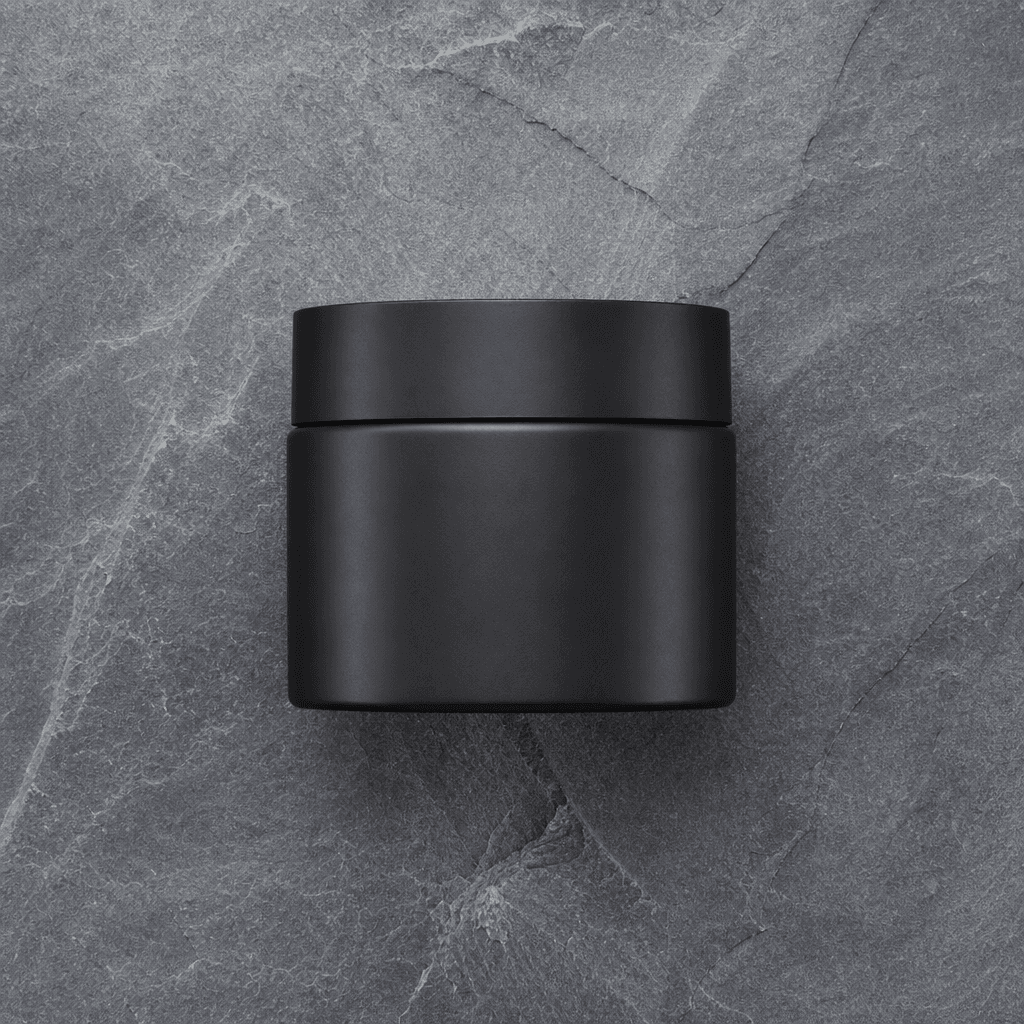 Overhead flat-lay of a matte black skincare moisturizer jar on a cool grey slate surface. Studio-perfect even lighting, 