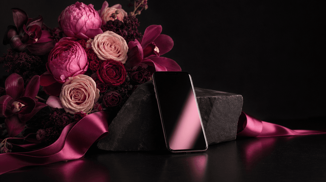 A carefully art-directed floral still life on a matte black set, bouquet, satin ribbon, dark stone, and a slim smartphon