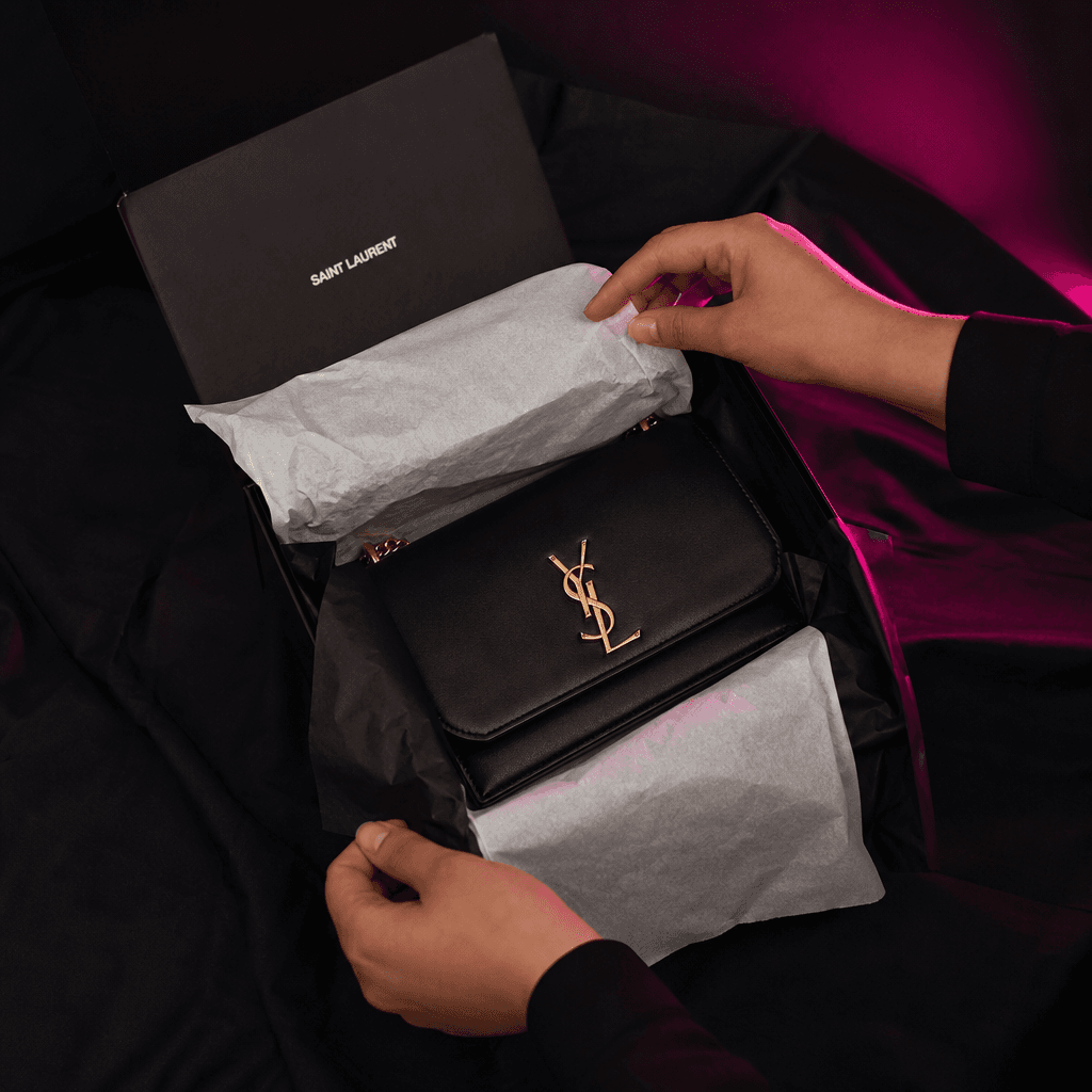 An elegant package being opened on a dark fabric surface, tissue paper unfolding around a carefully presented fashion ac