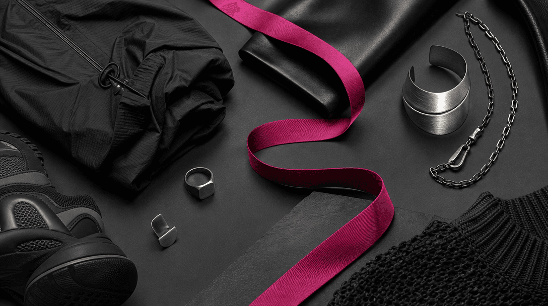 A curated arrangement of fashion materials on a dark studio floor: folded technical fabric, polished leather, brushed me