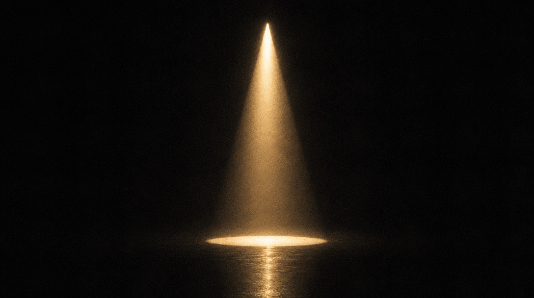A single tungsten beam of light descends through absolute darkness, casting a sharp perfect cone onto a reflective dark 