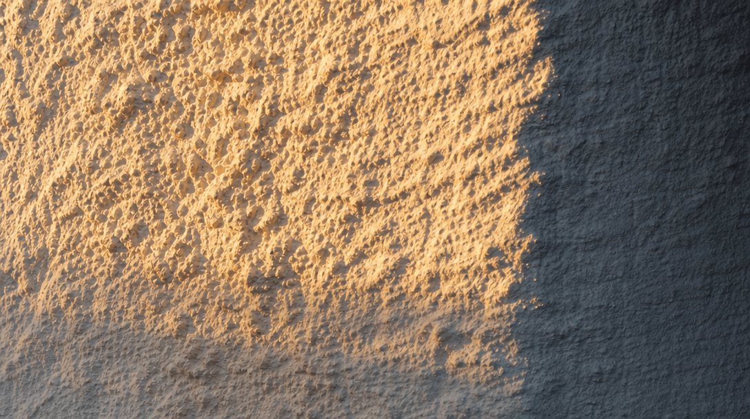 Extreme close-up of warm raking sunlight across a rough white plaster wall surface, grazing light revealing deep tactile