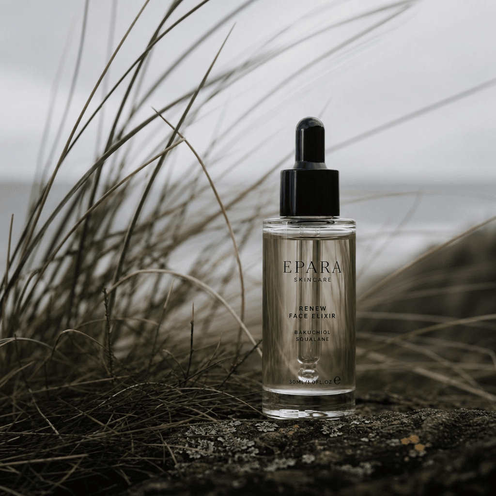 A small luxury skincare bottle standing among soft coastal grass, wind-shaped blades, overcast cinematic daylight, muted
