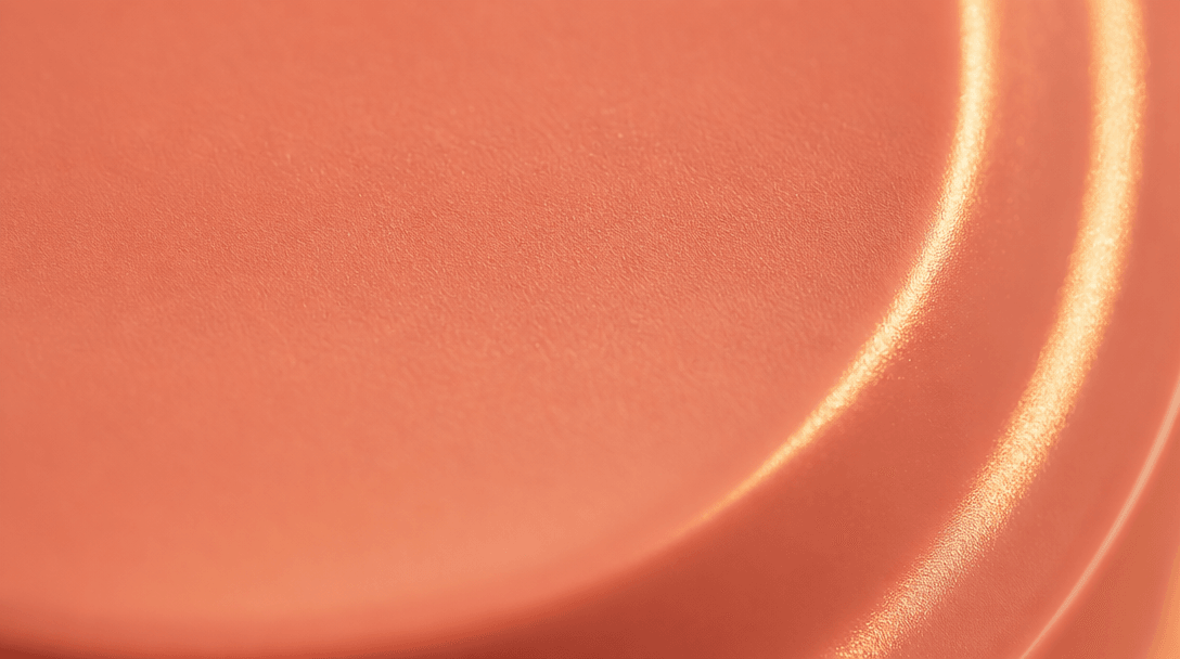 Extreme close-up of a smooth coral-pink cosmetic packaging surface under warm directional golden light. Subtle matte mat
