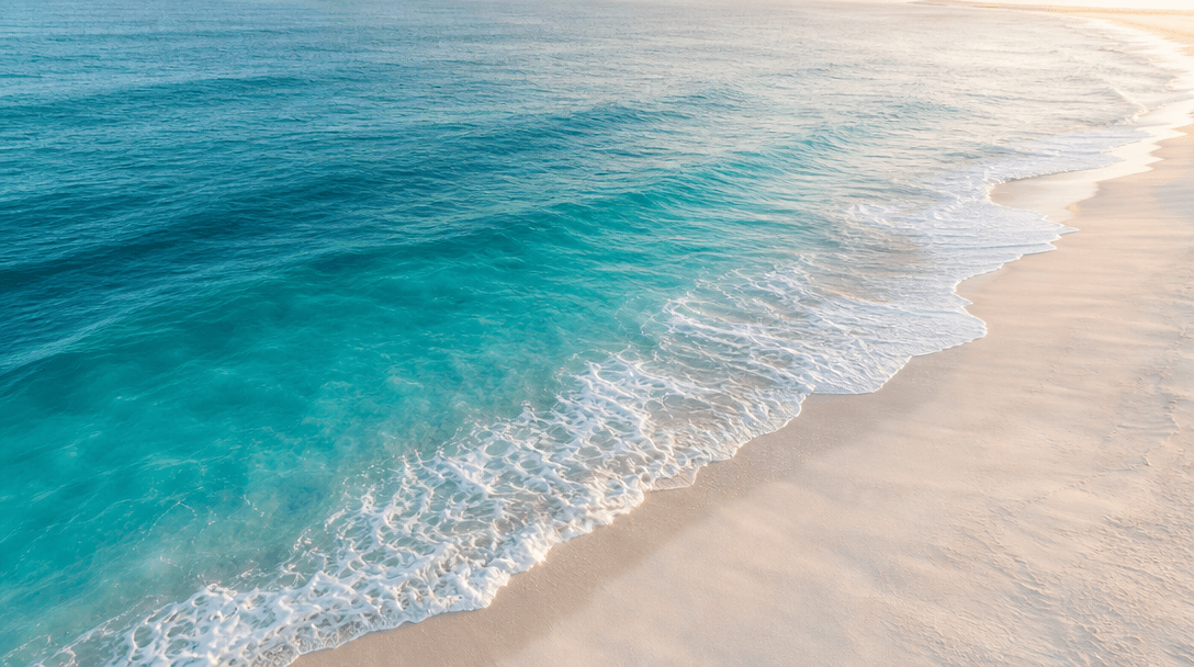 Atmospheric aerial-perspective view of ocean waves meeting white sand beach. Deep teal gradient water, white foam at the