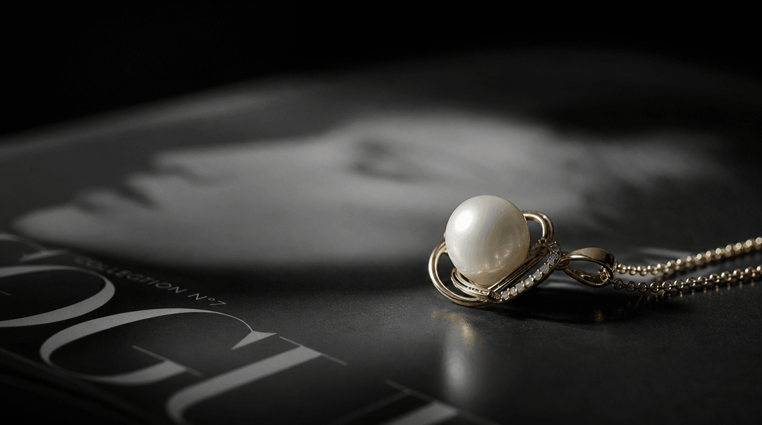 A pearl and gold pendant shot at a low editorial angle on a B&W print surface, controlled shallow depth of field with sh