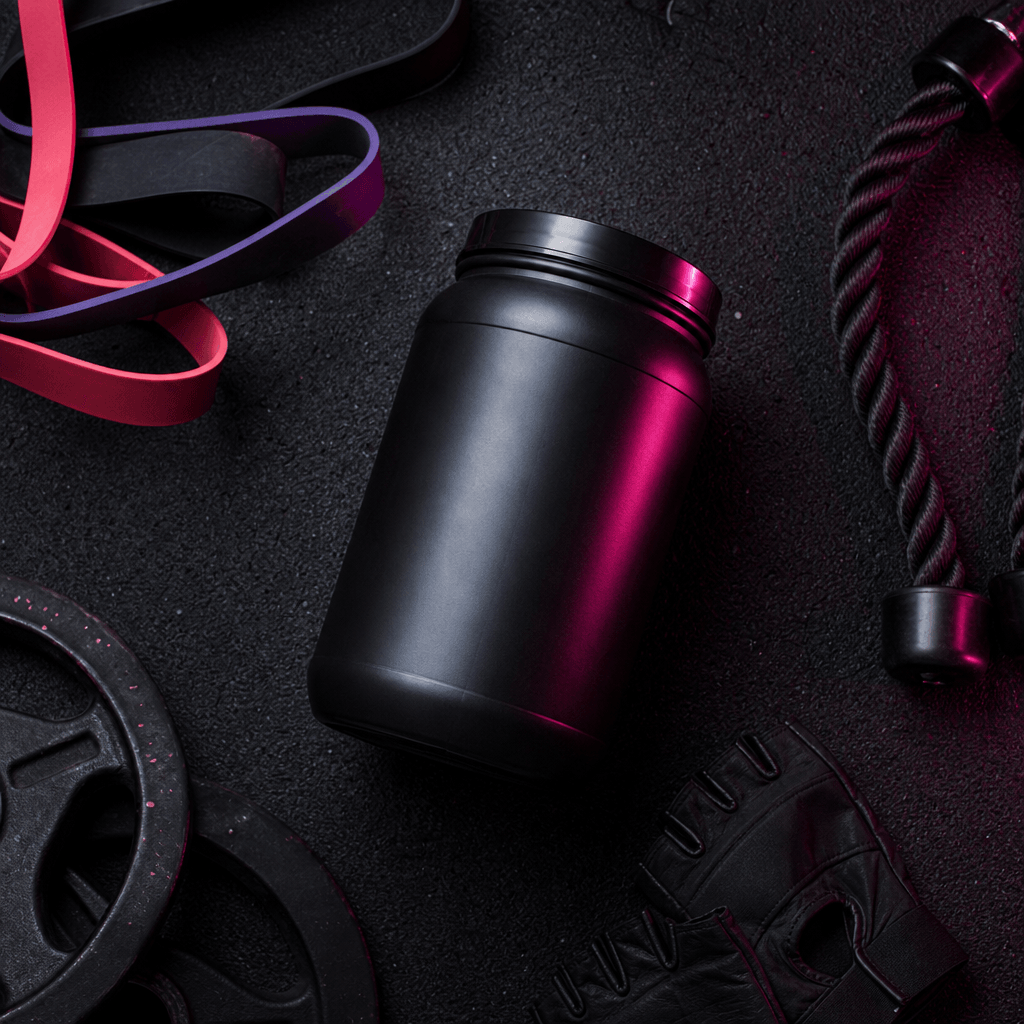 Overhead flat-lay of a premium sports supplement canister, resistance bands, and gym gloves arranged on black rubber flo