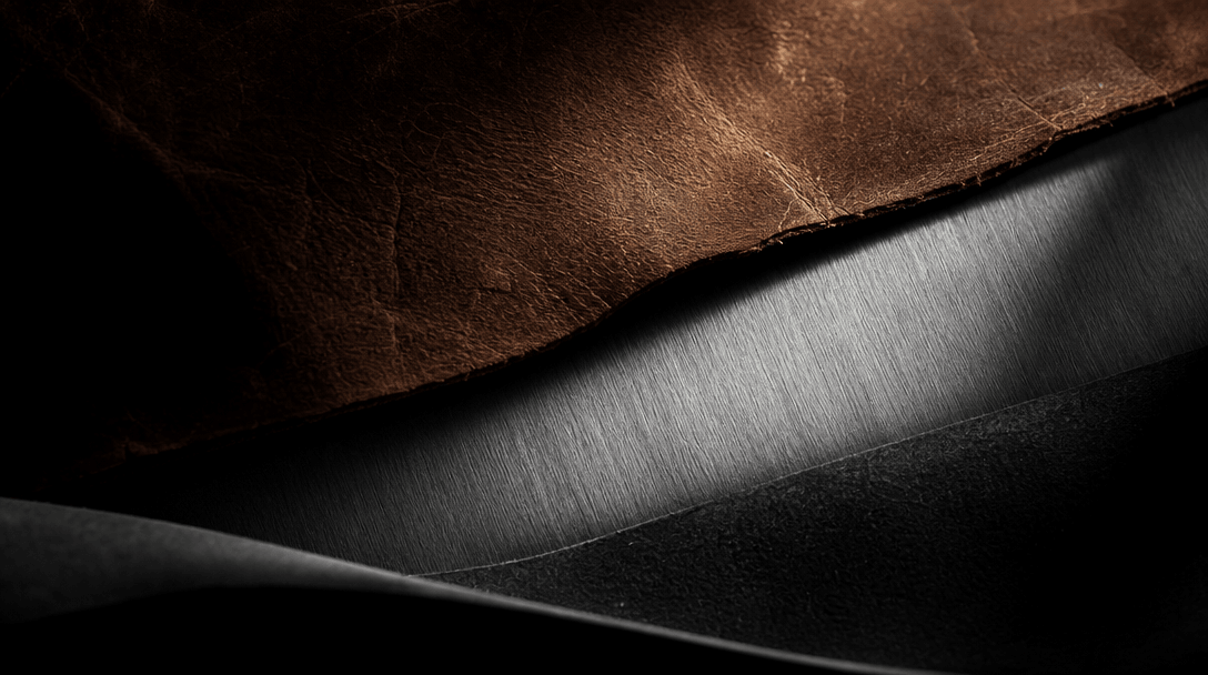 Abstract cinematic close-up of layered raw material textures — aged leather, brushed aluminum, matte black rubber — arra