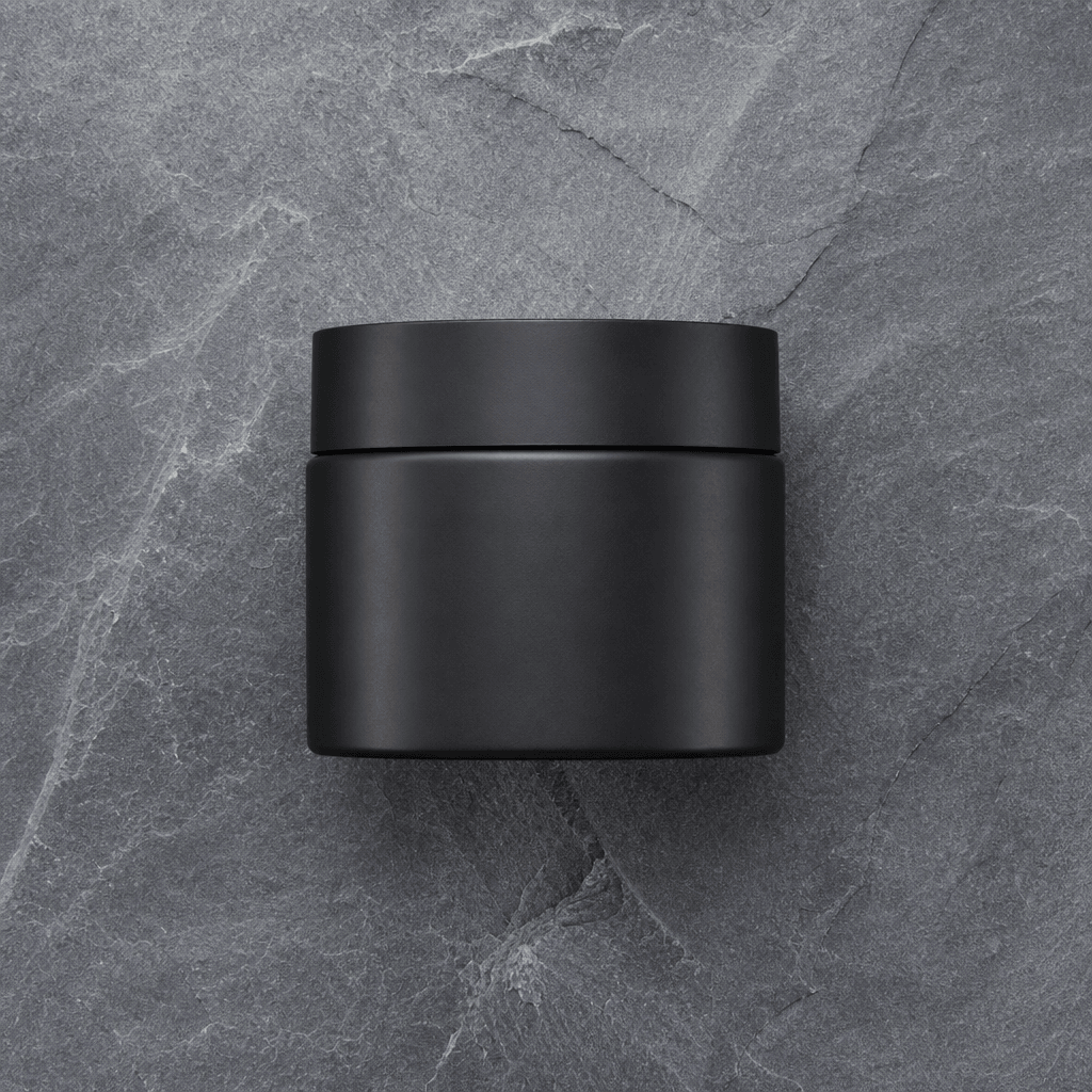 Overhead flat-lay of a matte black skincare moisturizer jar on a cool grey slate surface. Studio-perfect even lighting, 