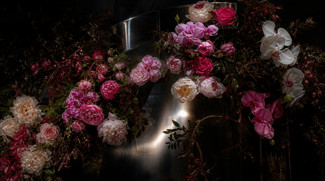 A dense landscape of roses, peonies, orchids, and glossy leaves sweeping across dark reflective metal like a floral inst
