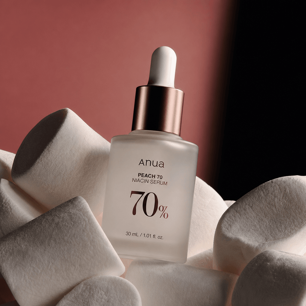 A frosted glass facial serum bottle nestled between oversized ivory marshmallows, soft blush background fading into deep