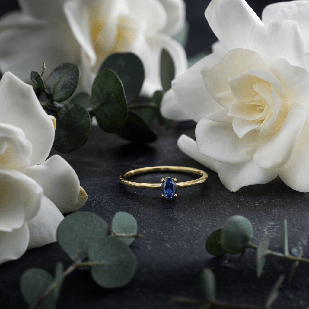 A slender gold ring set with a small oval sapphire nestled between white gardenia blooms and eucalyptus sprigs on a dark