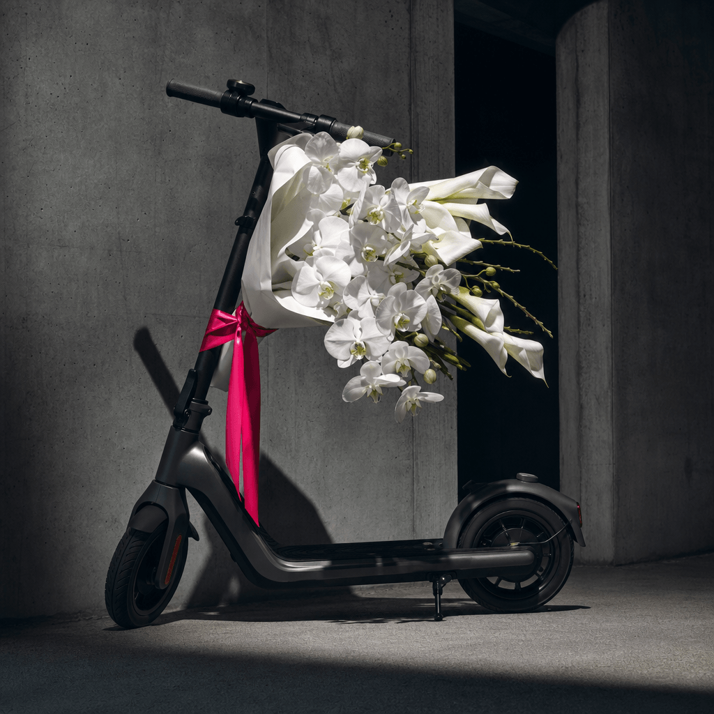 A sculptural white orchid and calla lily bouquet transported on a sleek charcoal electric scooter, modern concrete archi