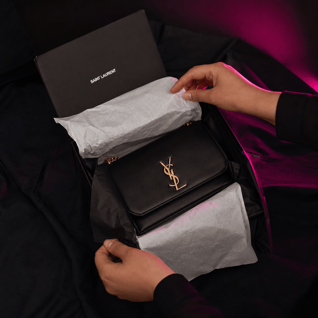 An elegant package being opened on a dark fabric surface, tissue paper unfolding around a carefully presented fashion ac