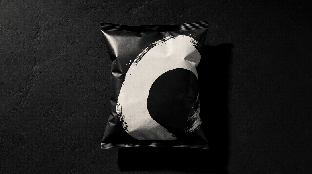 Dark editorial flat-lay: a single sealed snack product with bold graphic packaging centered on a deep near-black charcoa