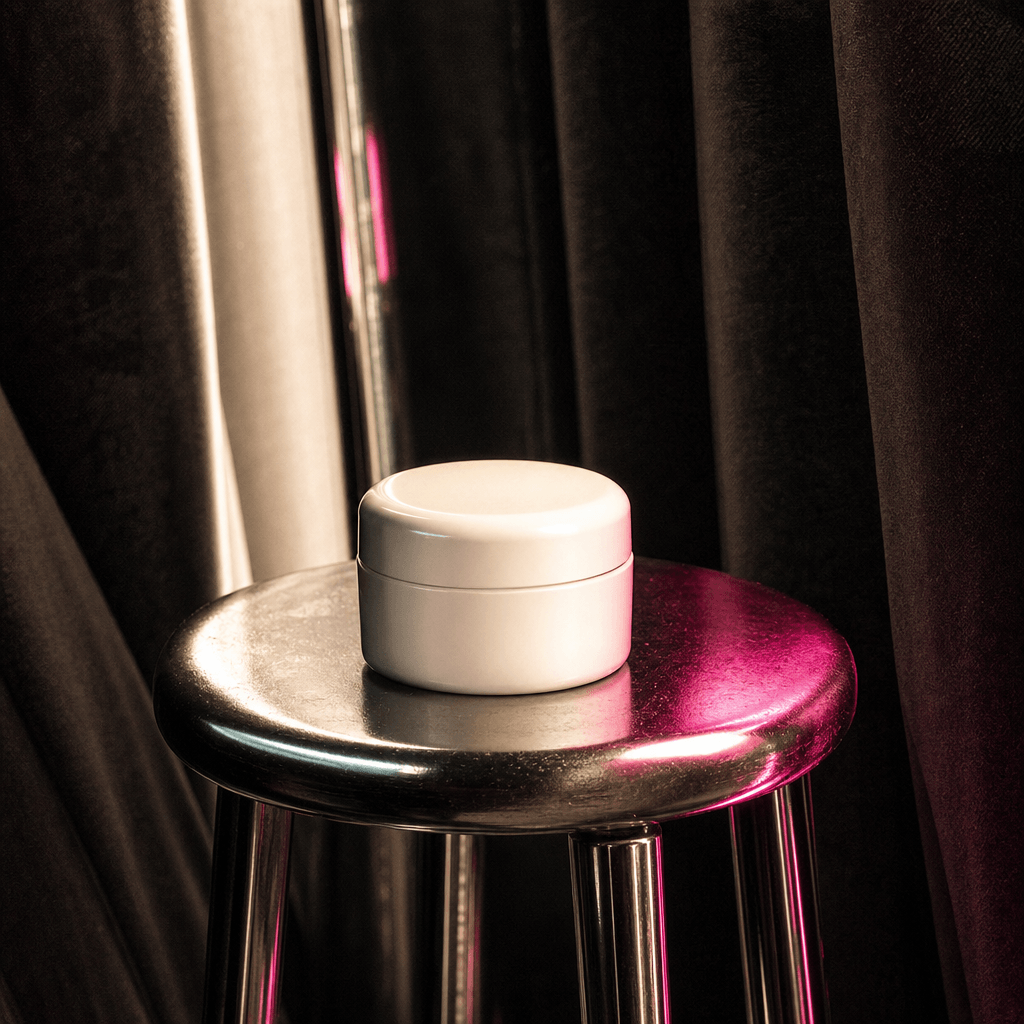 Minimal skincare jar on a chrome stool within a dark photobooth, surrounded by soft curtain folds and rhythmic flash ref