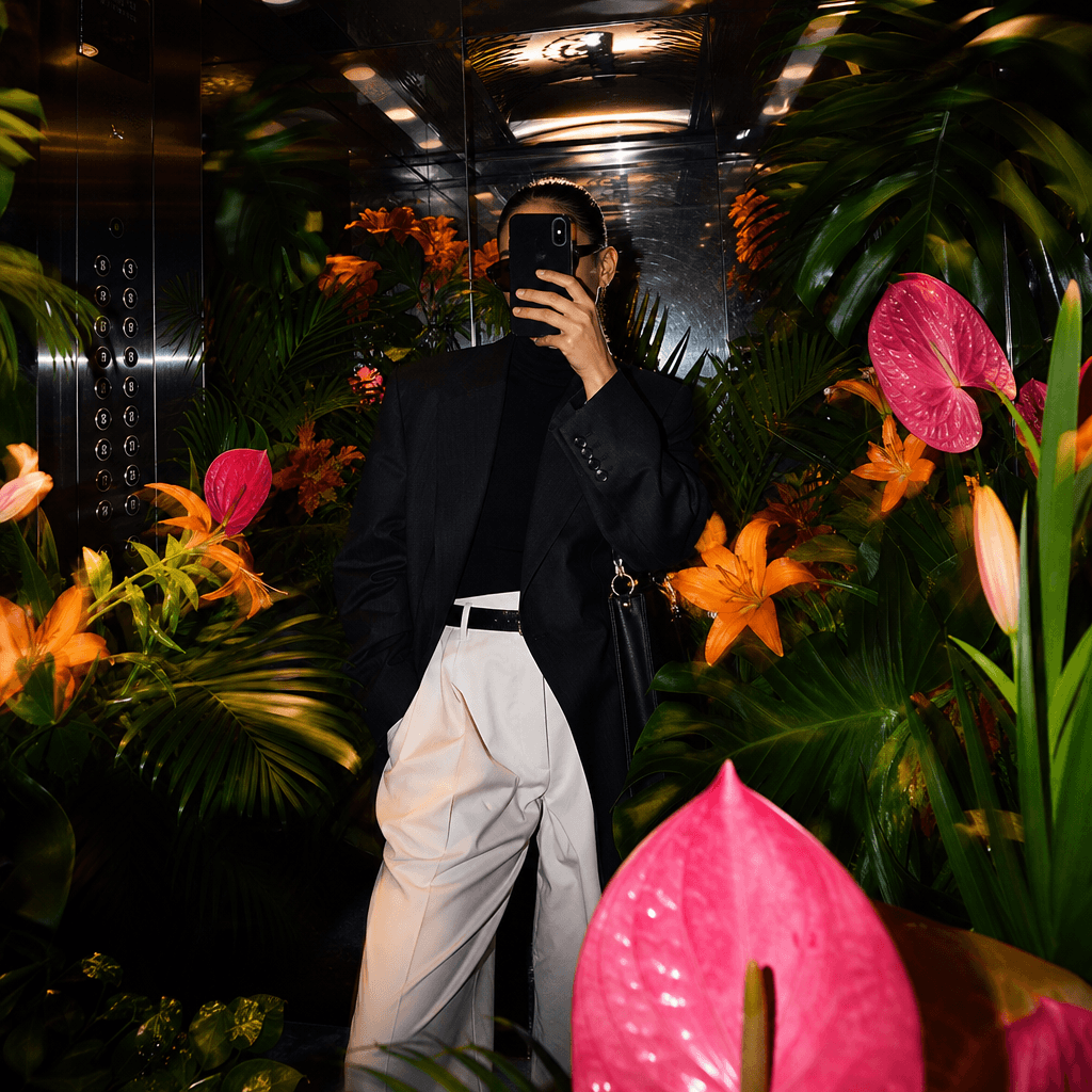 A maximalist fashion mirror selfie in an elevator transformed by vivid orange lilies, magenta anthuriums, and oversized 