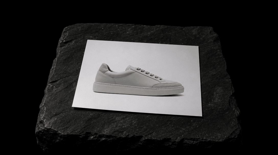 Dark editorial still life: a single sharp product photograph — a minimalist sneaker on a clean white surface — printed a
