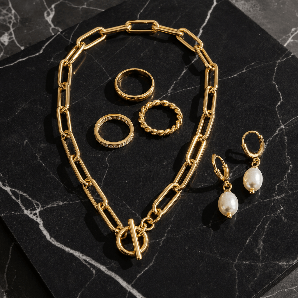 Flat lay of a luxury jewelry set — gold link chain necklace, stacking rings, and pearl drop earrings — arranged on polis