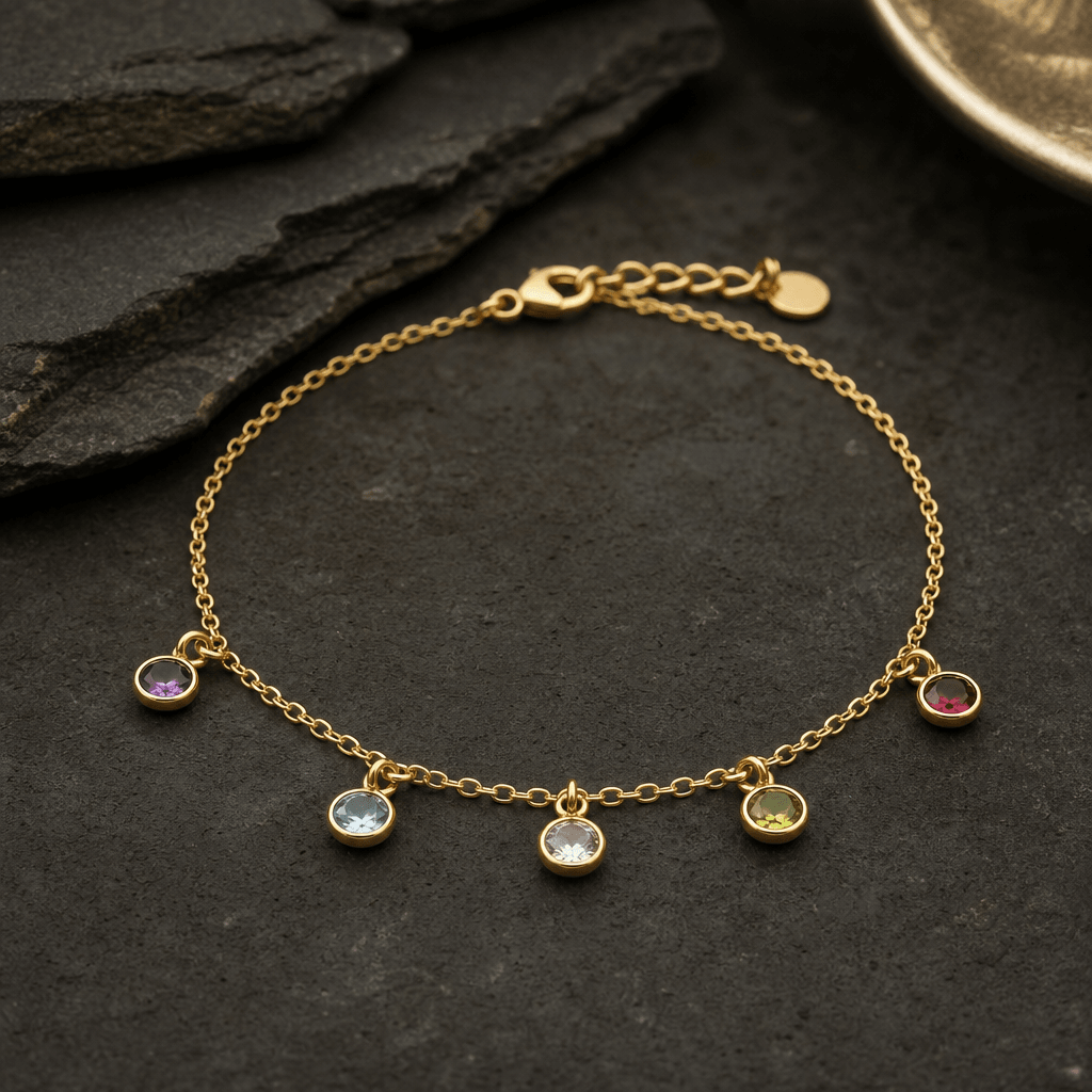 Square studio product shot: a delicate gold chain bracelet with small gemstone pendants laid flat on a matte dark stone 
