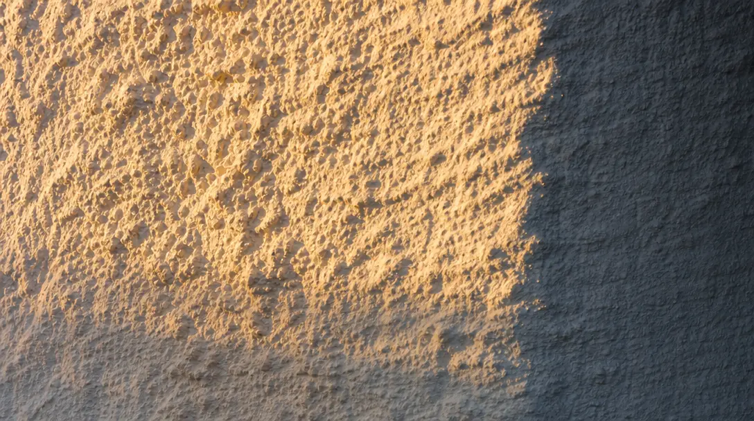 Extreme close-up of warm raking sunlight across a rough white plaster wall surface, grazing light revealing deep tactile