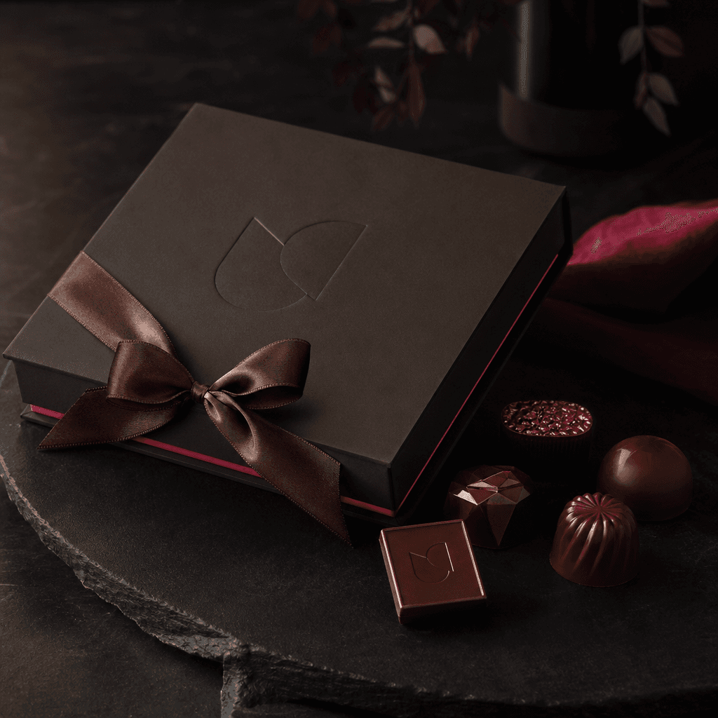 Elegant corporate gifting scene with a closed charcoal chocolate box, embossed abstract logo, satin ribbon, and several