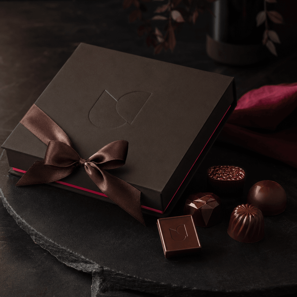 Elegant corporate gifting scene with a closed charcoal chocolate box, embossed abstract logo, satin ribbon, and several