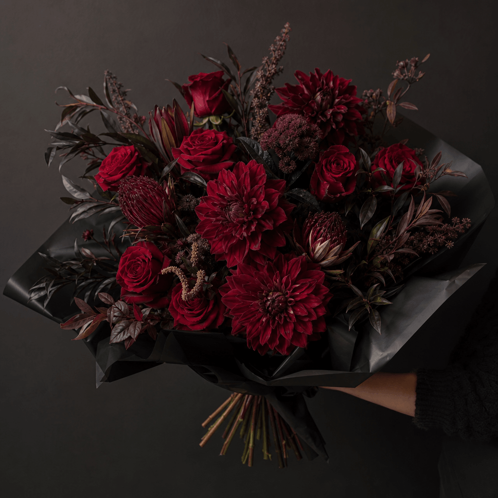 A dramatic bouquet of burgundy dahlias, deep red roses, and dark foliage held against a charcoal plain wall, sculptural