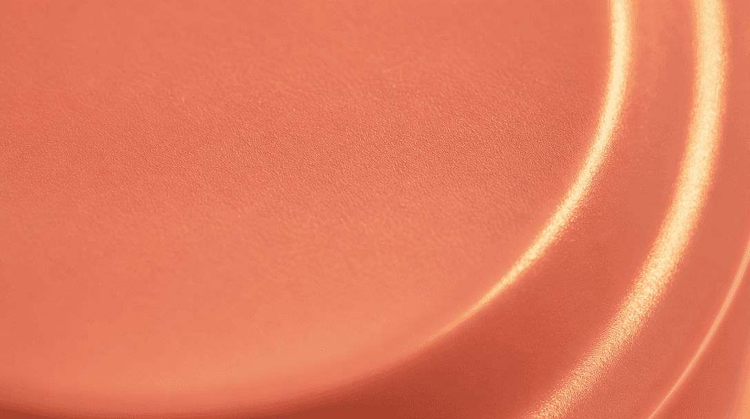 Extreme close-up of a smooth coral-pink cosmetic packaging surface under warm directional golden light. Subtle matte mat