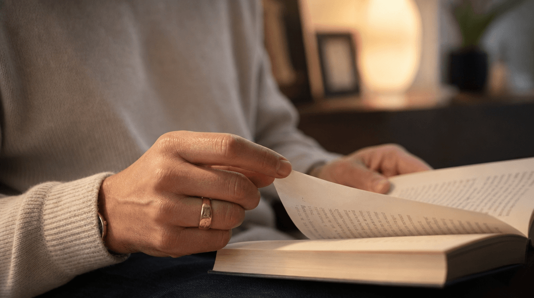 Close editorial framing of hands mid-activity: a book open, fingers caught mid-page-turn, warm lamp glow catching paper