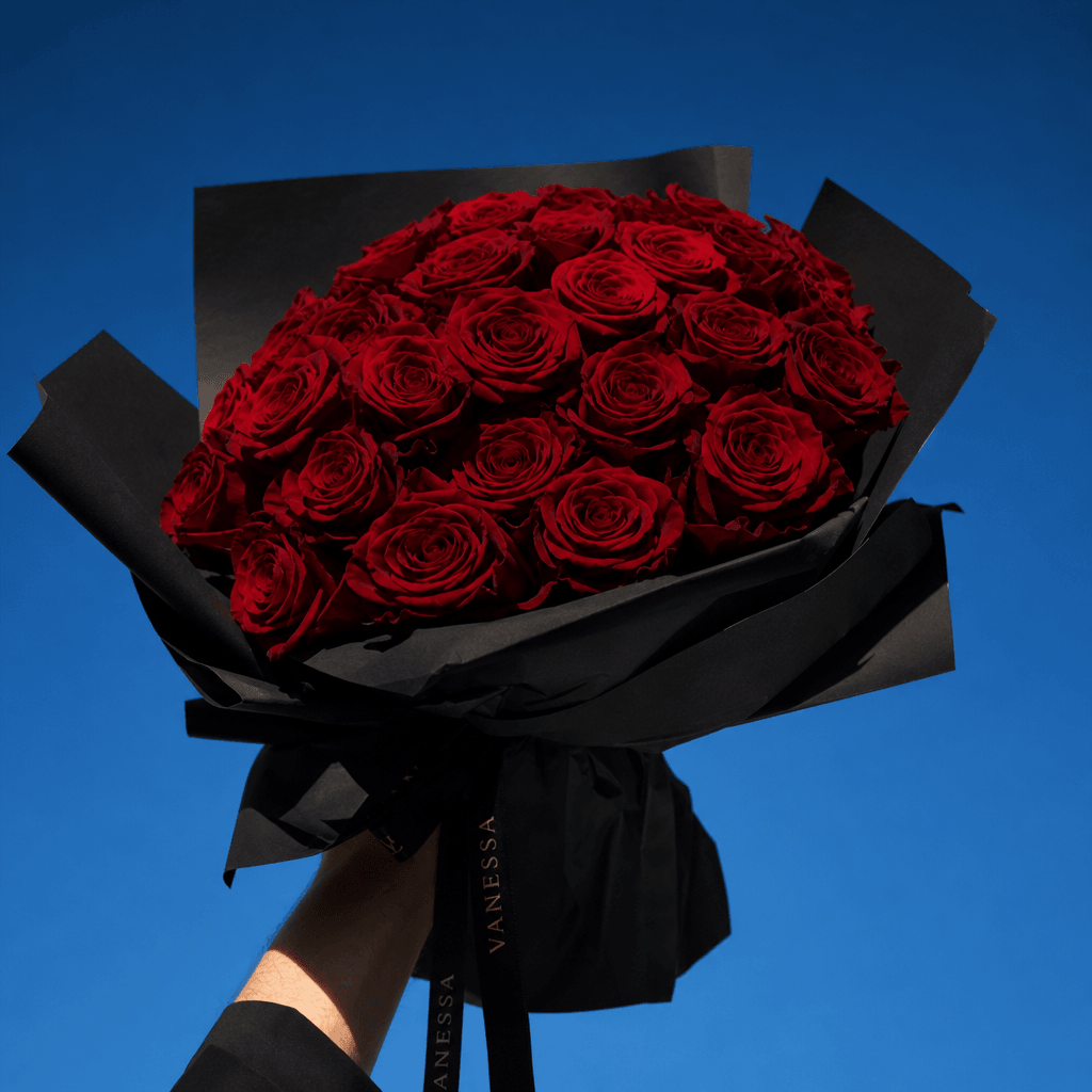 A sculptural bouquet of deep red roses wrapped in matte black paper, lifted against a cloudless cobalt sky, cinematic lo