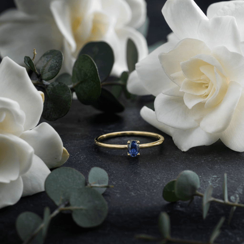 A slender gold ring set with a small oval sapphire nestled between white gardenia blooms and eucalyptus sprigs on a dark
