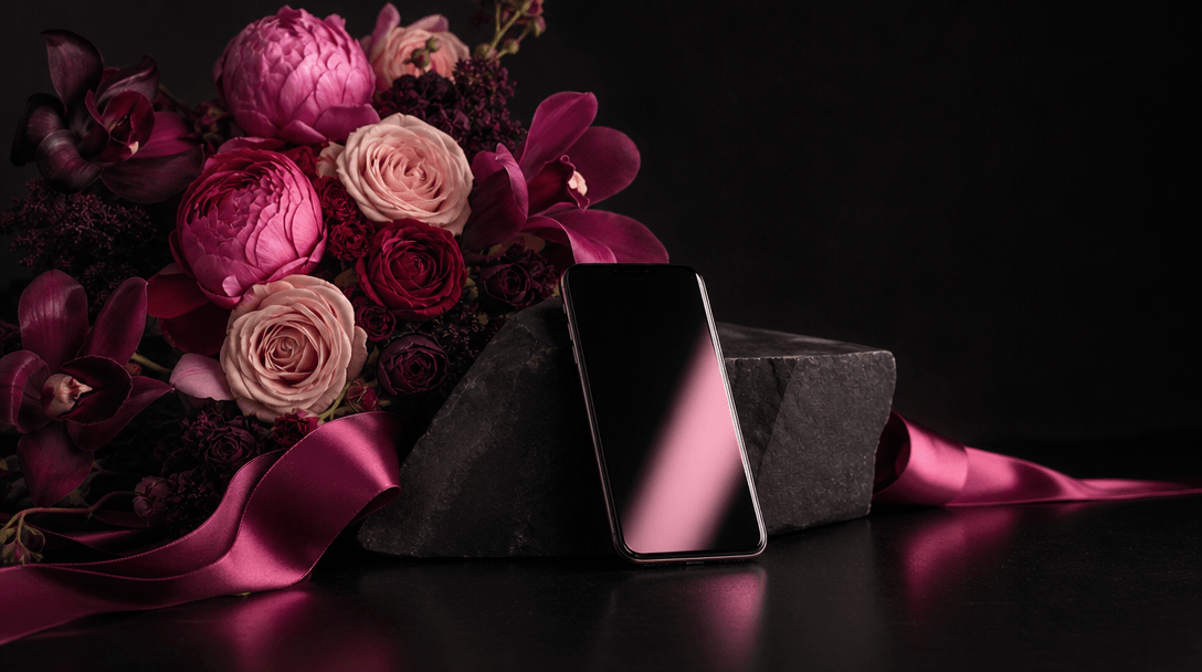 A carefully art-directed floral still life on a matte black set, bouquet, satin ribbon, dark stone, and a slim smartphon