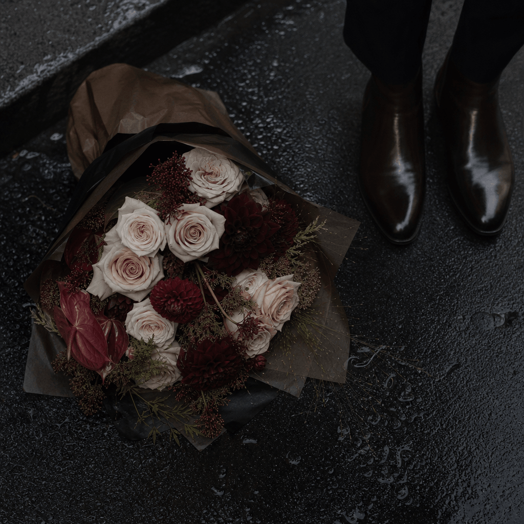 A loose bouquet of pale roses and deep burgundy flowers placed on rain-darkened asphalt beside polished ankle boots. Ove