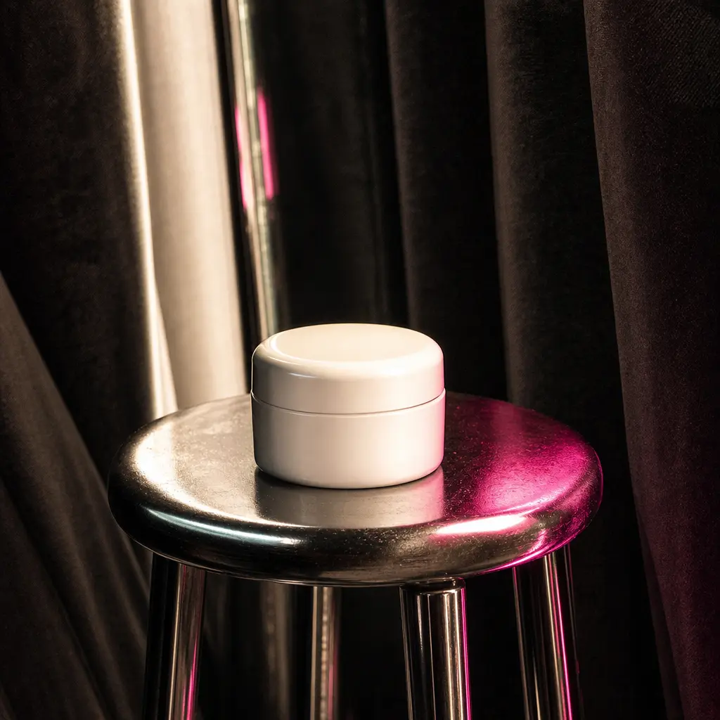 Minimal skincare jar on a chrome stool within a dark photobooth, surrounded by soft curtain folds and rhythmic flash ref