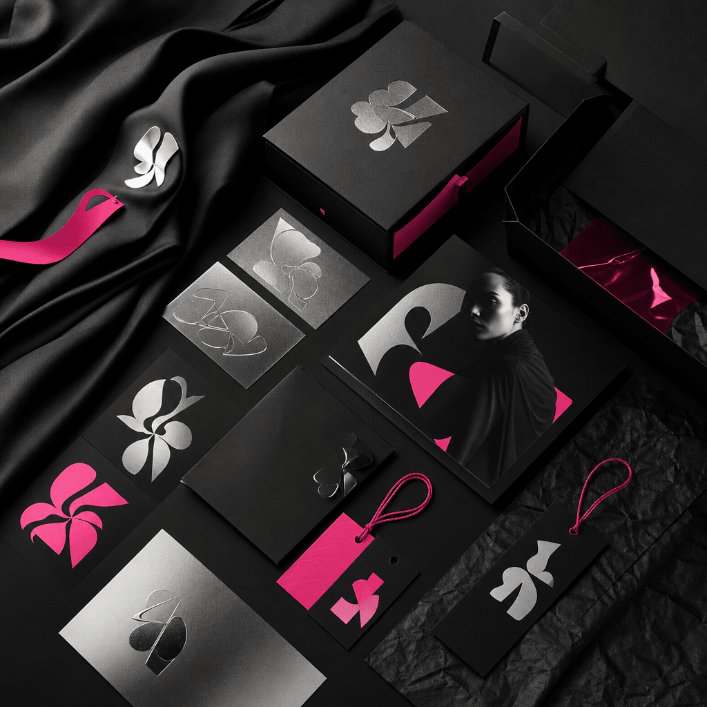 A modern fashion label identity board with folded black fabric, silver foil cards, vivid hot pink accents, abstract typo