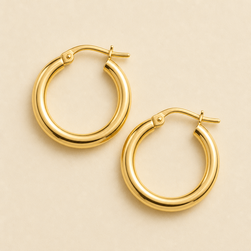 Overhead studio close-up of a pair of yellow gold hoop earrings arranged flat on a warm matte ivory surface. Softbox lig