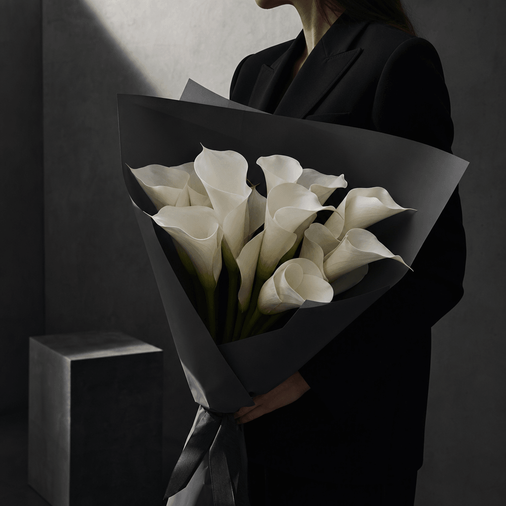 A modern white calla lily bouquet wrapped in matte charcoal paper, carried by an anonymous model wearing a sharply tailo