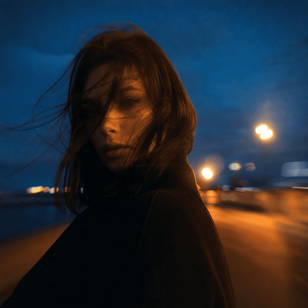 Square outdoor portrait of an anonymous creative at blue hour, windswept coat, distant sodium streetlights, deep navy sk