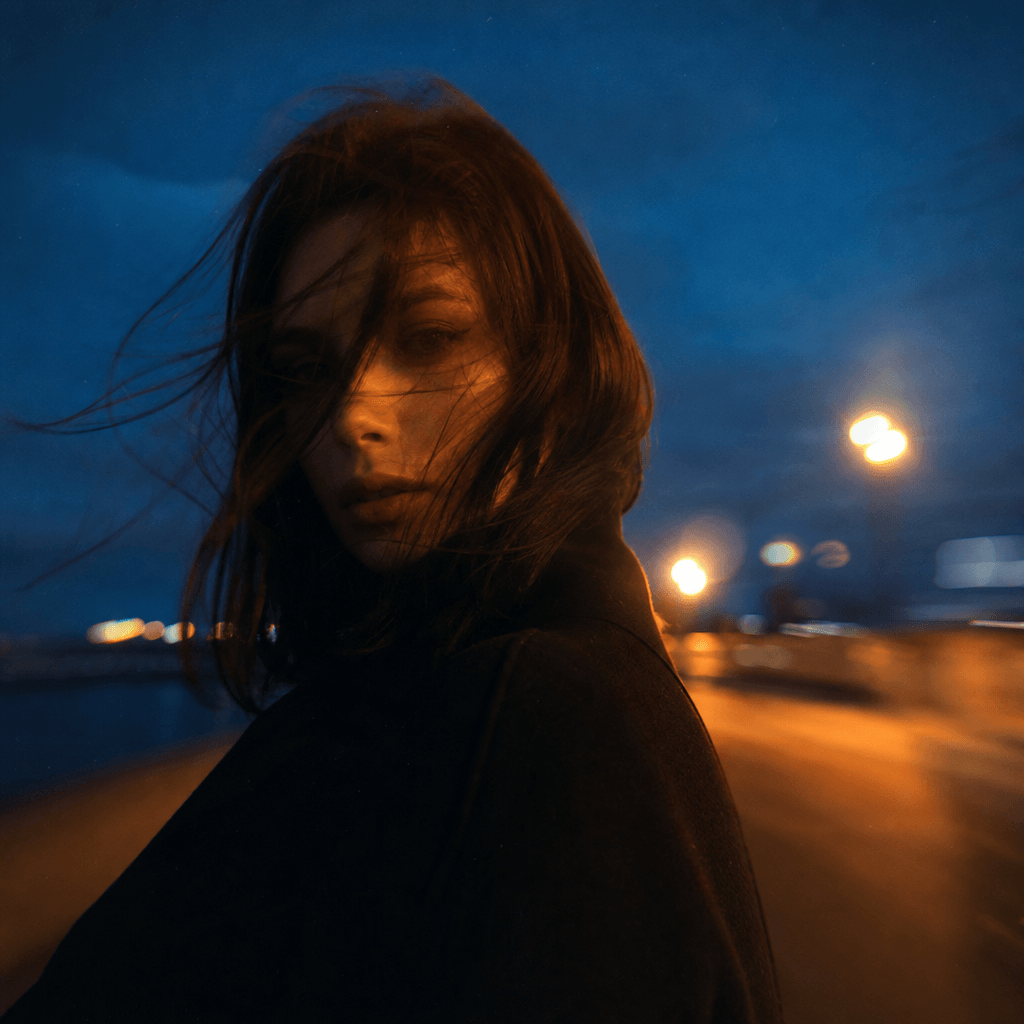 Square outdoor portrait of an anonymous creative at blue hour, windswept coat, distant sodium streetlights, deep navy sk